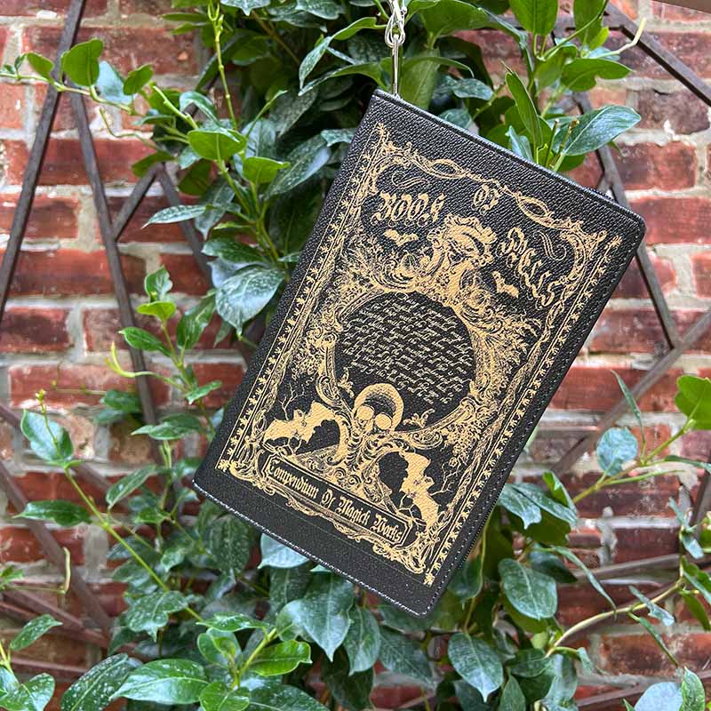 Black and gold book-themed pouch hanging from green leaves against a brick wall.