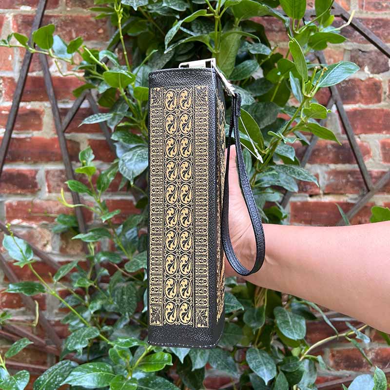 Hand holding a black clutch with gold patterns against a brick wall and green plants background