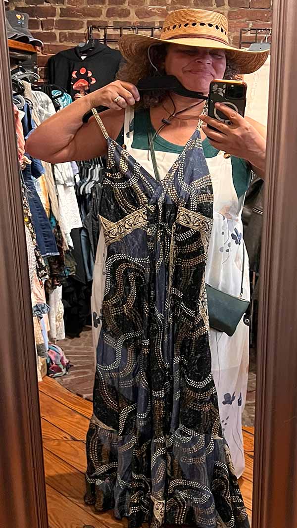 Woman taking a mirror selfie wearing the midnight blue and rich blue colors in this version of the boho maxi dress with adjustable straps that is available in on size fits 0-10, while wearing a straw hat in a clothing store.