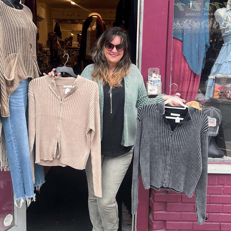 model standing in doorway, wearing green double zip sweater as a cardigan, holding tan double zip sweater on a hanger in one hand , and grey double zip sweater in the other hand