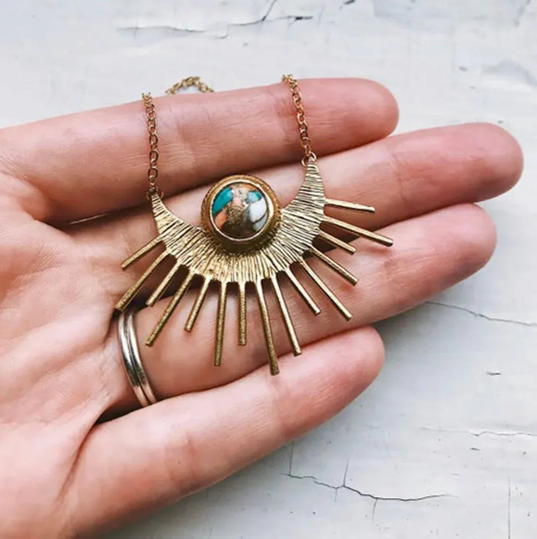 Gold-tone sunburst pendant necklace with teal center; layered rays on a thin chain