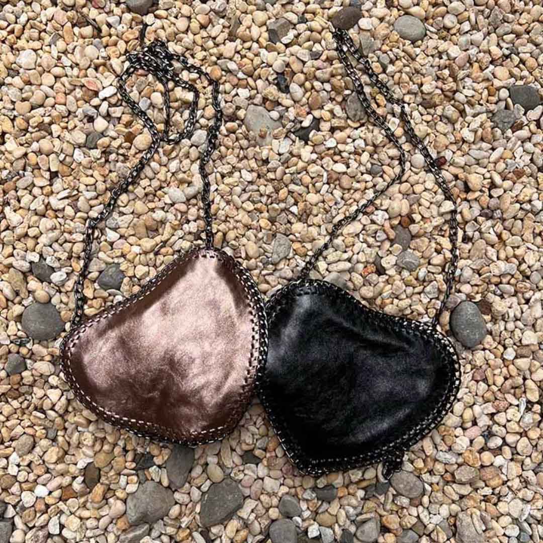 One bronze and one black heart shape purse with crossbody chain laying in a bed of stones