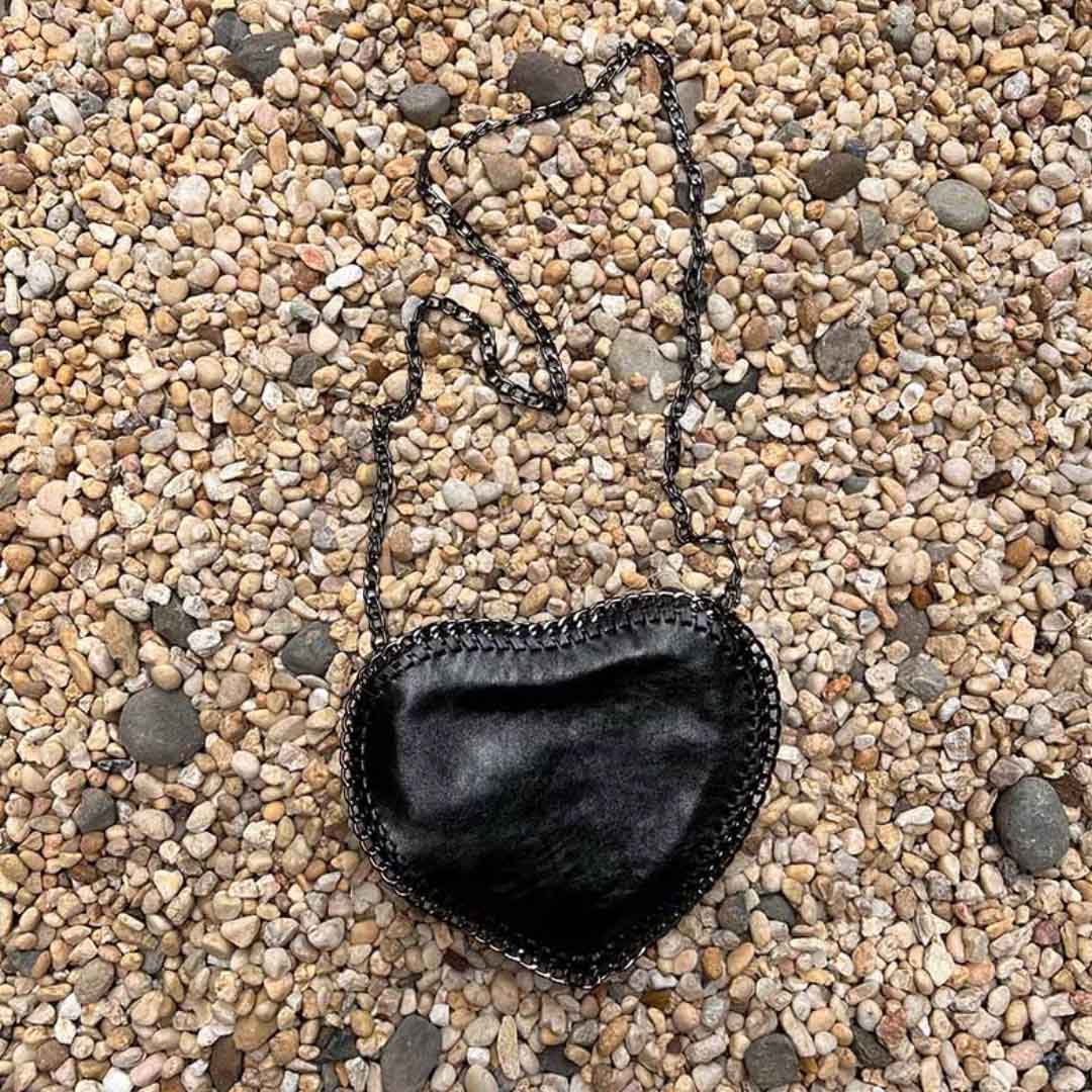 Black heart shape purse with crossbody chain laying in a bed of stones