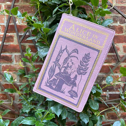 Purple Alice in Wonderland book purse is hanging in the secret garden on green foliage with a brick wall background