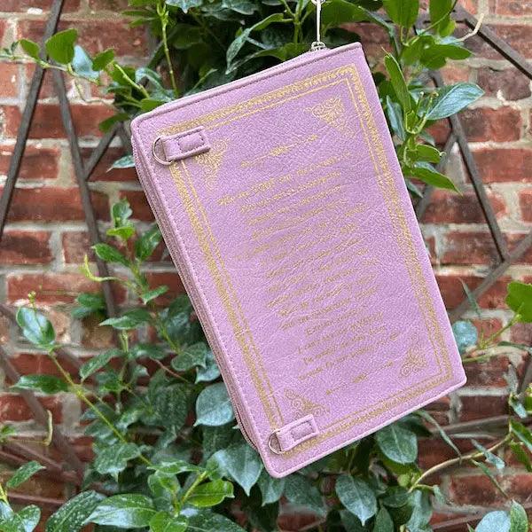 Purple book-shaped pouch with gold text and design, hanging from green leaves against a brick wall.