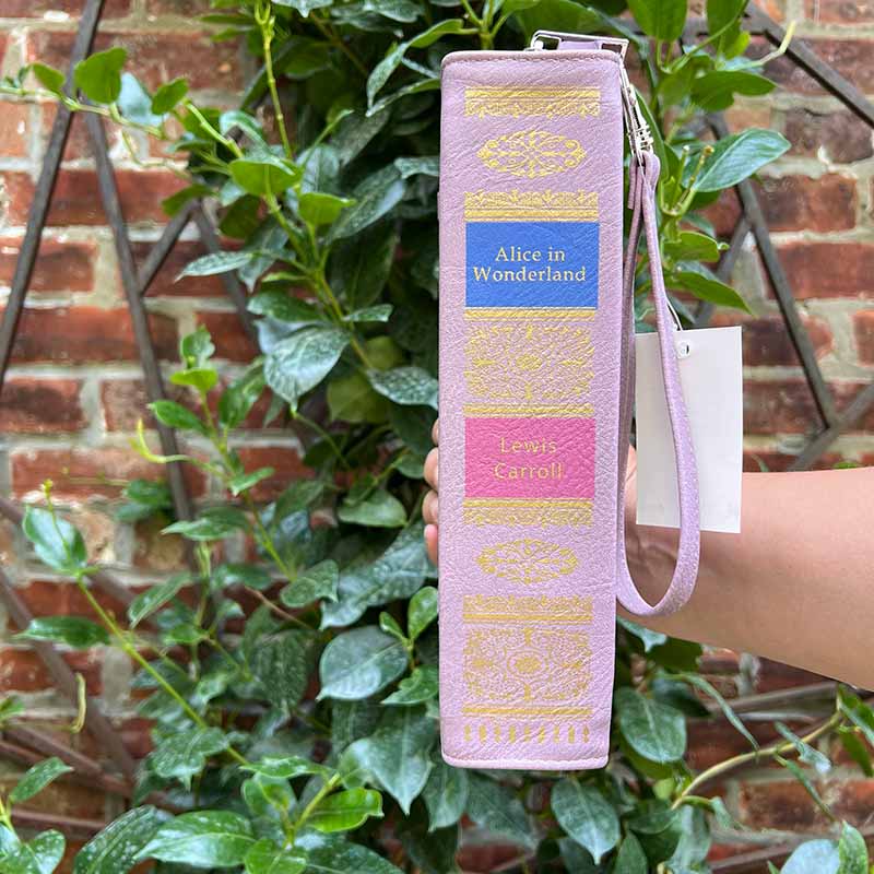 Purple book-shaped bag with gold detailing and a blue label. The Spine of the Alice in Wonderland Storybook handbag is held in front of a mandavilla.