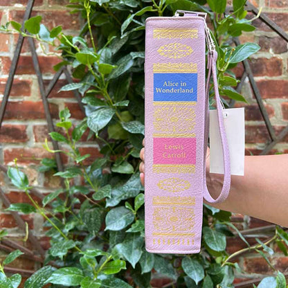 Purple book-shaped bag with gold detailing and a blue label. The Spine of the Alice in Wonderland Storybook handbag is held in front of a mandavilla.