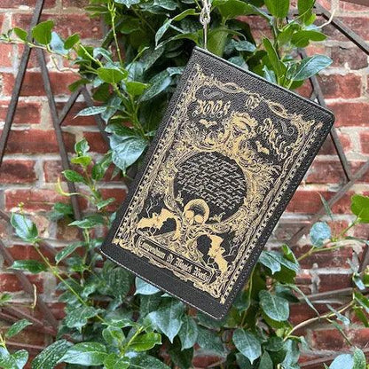 Decorative Book of Spells handbag  with gold embossed  witchy, ethereal designs hanging from greenery against a brick wall.