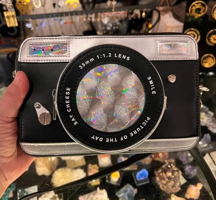 Camera crossbody bag front view, large circular lens graphic, silver edges, side buckles