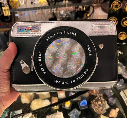 Camera crossbody bag front view, large circular lens graphic, silver edges, side buckles