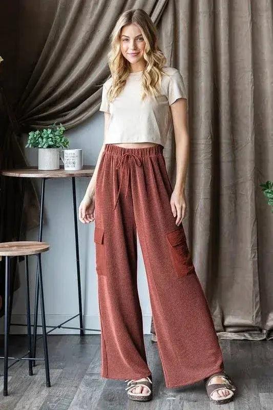 Woman wearing a white top and rust-colored wide-leg rust cargo pants standing in a room with curtains and a plant. Elastic waistband and drawstring detail.