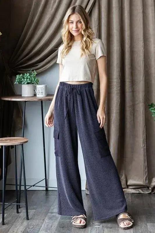 Woman wearing a white top and -wide-leg navy ribbed cargo pants standing in a room with curtains and a plant. Elastic waistband and drawstring detail.