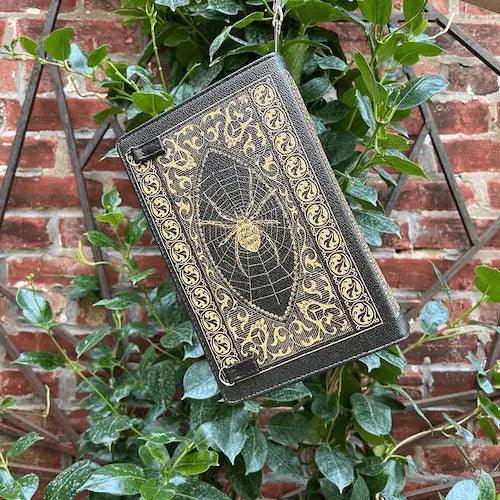 Decorative Dracula book shaped handba with intricate gold  spiderweb and black window designs hanging from mandavilla leaves against a brick wall in the secret garden.