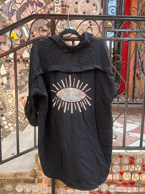 Black linen shirt with a decorative eye design on a hanger against a mosaic wall.