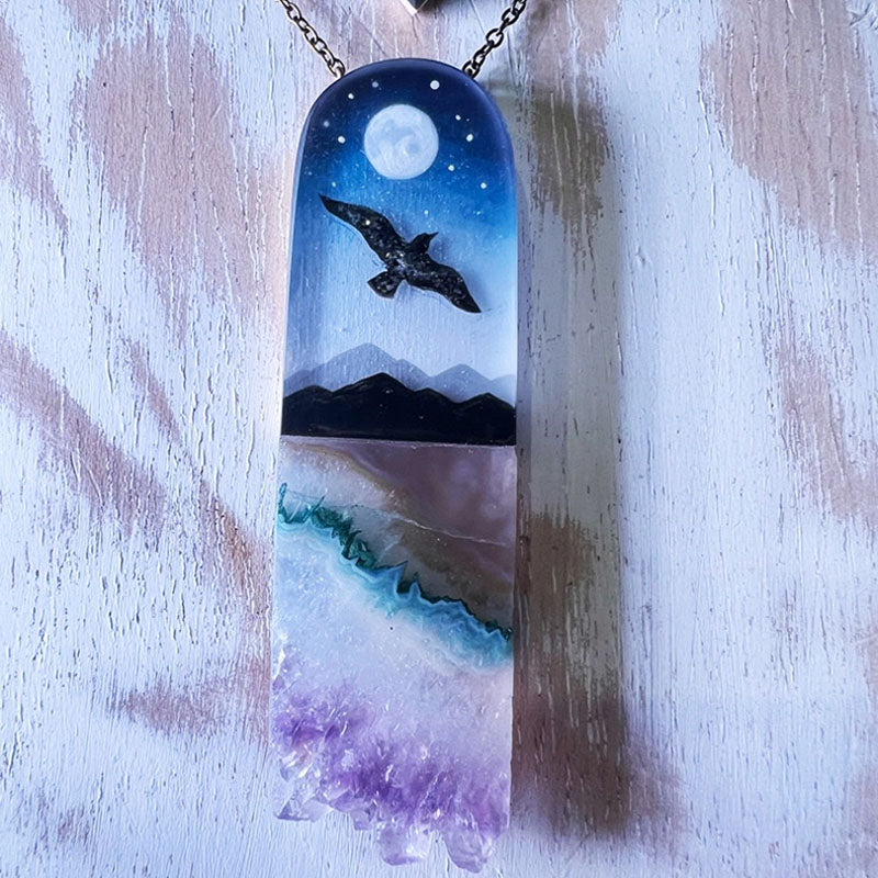 Necklace with a pendant featuring a night sky design with a bird, moon, and mountains on a wooden surface.
