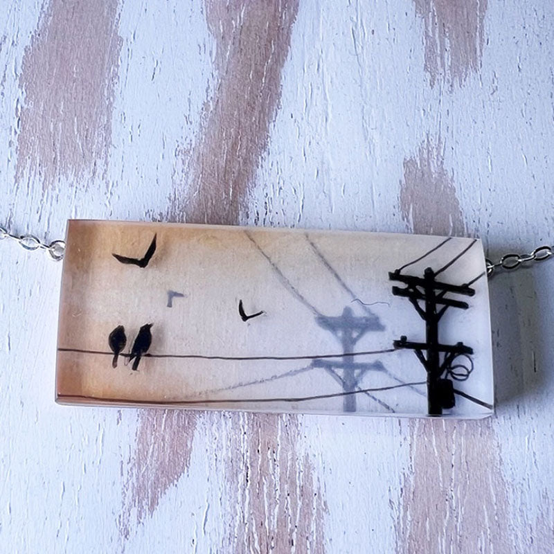 Necklace with pendant featuring birds on a wire and a telephone pole against a textured background