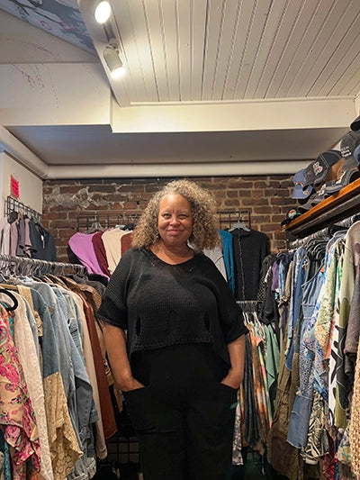 A woman with curly gray hair stands in a clothing boutique, wearing a black short-sleeve top and black pants. Clothing racks are visible on both sides of the aisle, and a brick wall is behind her.