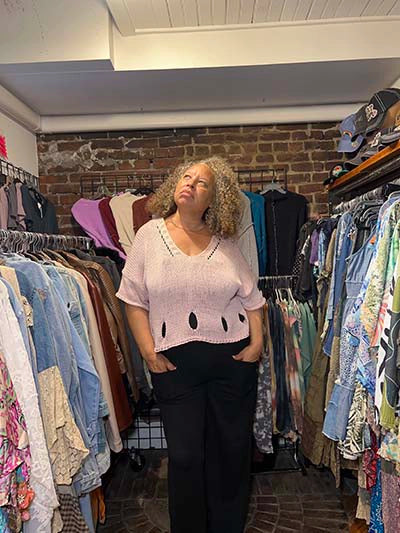 A woman with curly hair stands in a clothing boutique wearing a light pink knit top with black pants, surrounded by racks of clothes.