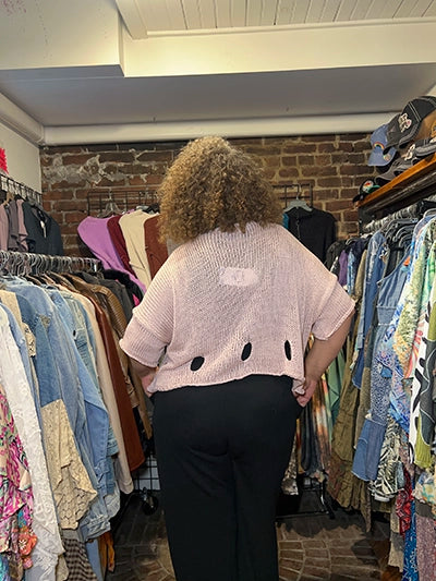 A person viewed from behind wearing a light pink short-sleeve knit top with three black oval cutouts and black pants, standing in a clothing store aisle surrounded by racks of garments.