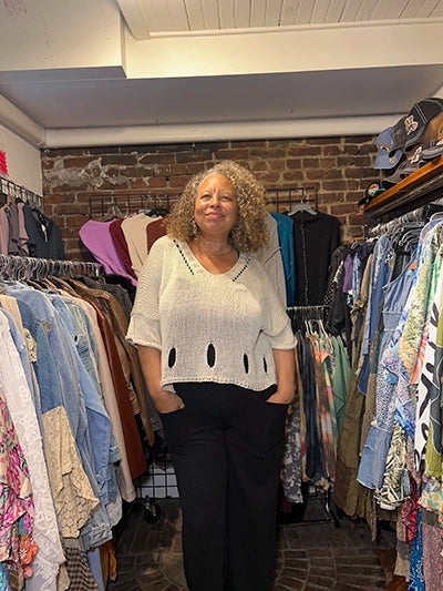 A woman with curly hair stands in a clothing boutique wearing a cream-colored knit sweater with cutout details and black pants, surrounded by racks of clothes.