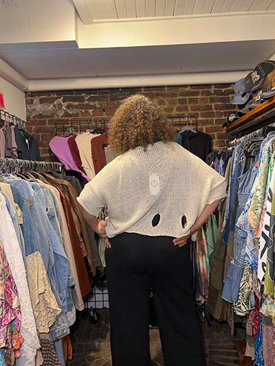 A person viewed from behind wearing a cream-colored, short-sleeved knit top with circular cutouts at the lower back, paired with black wide-leg pants, standing inside a clothing boutique surrounded by racks of garments.