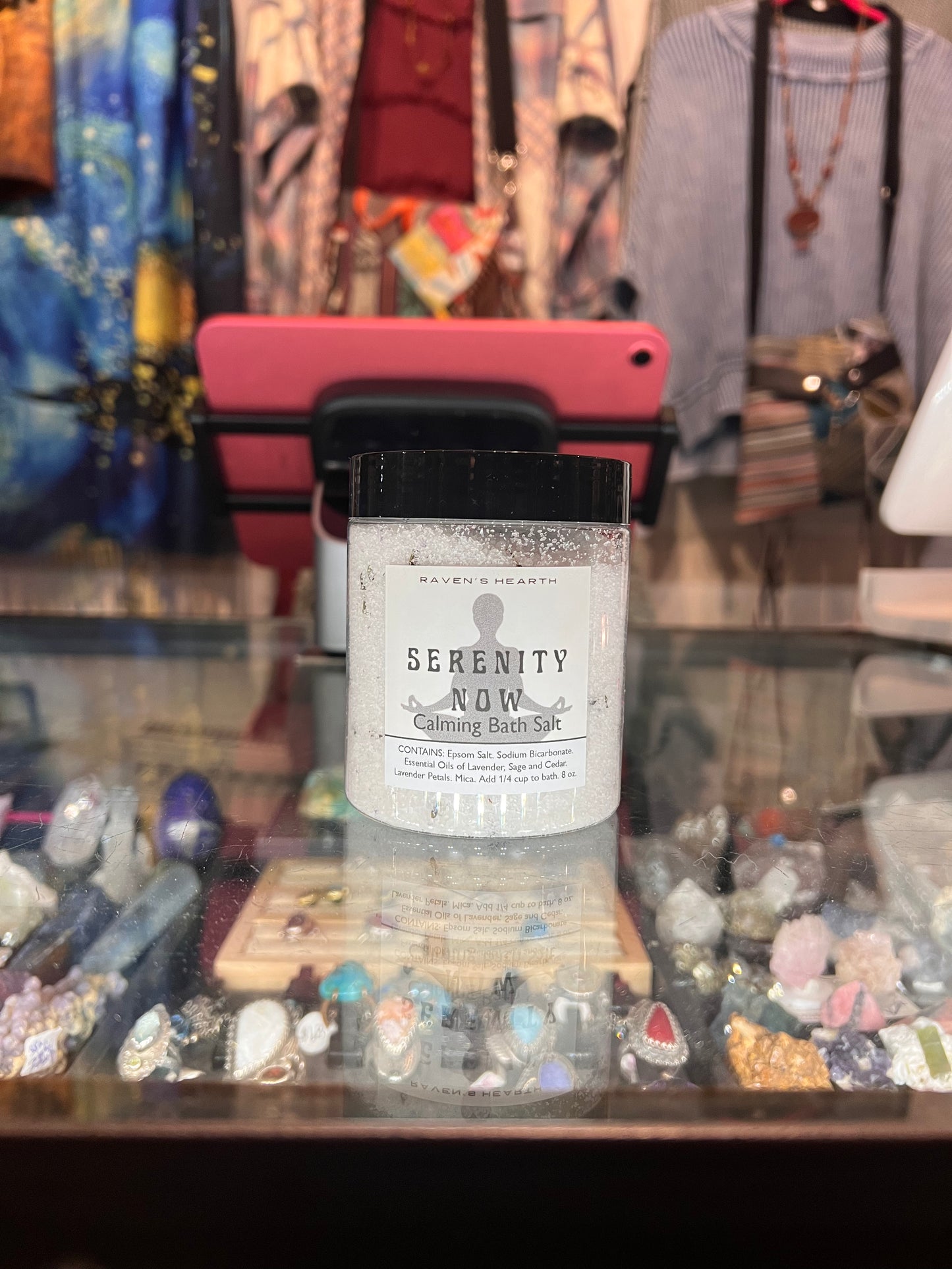 Bottle of 'Serenity Now' balm in a store setting with jewelry and clothing in the background