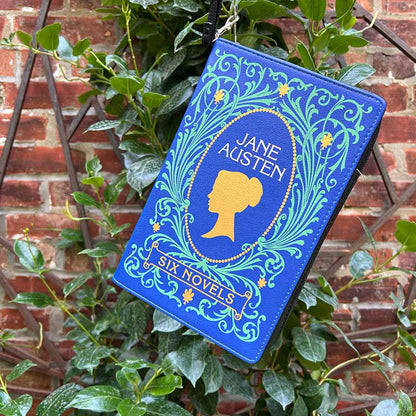 Blue clutch and crossbody handbag with Jane Austen design hanging on green leaves against a brick wall.