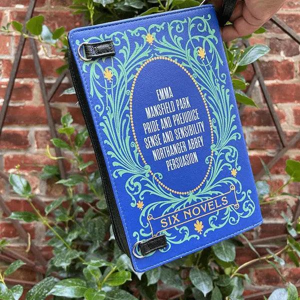 Blue storybook purs featuring the 6 novels of jane austen with literary design against a brick wall and plant background