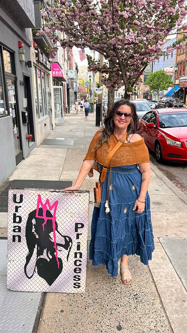 Dark denim boho maxi dress styled for a city stroll, standing next to Urban Princess sign with a cross body bag and summer crochet shawl