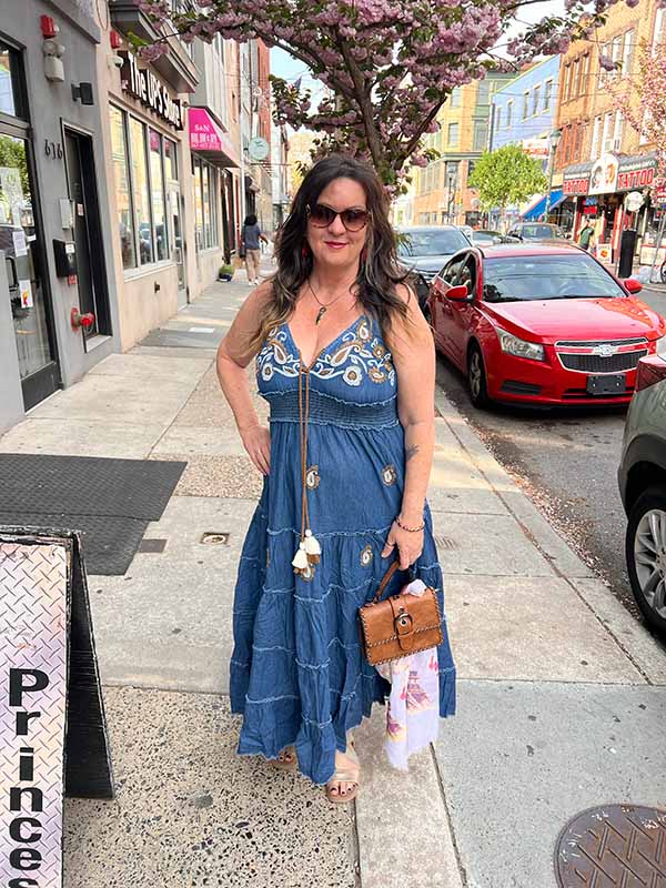 Owner Mary wearing a dark denim beaded maxi dress with adjustable straps on a city street with cherry blossoms