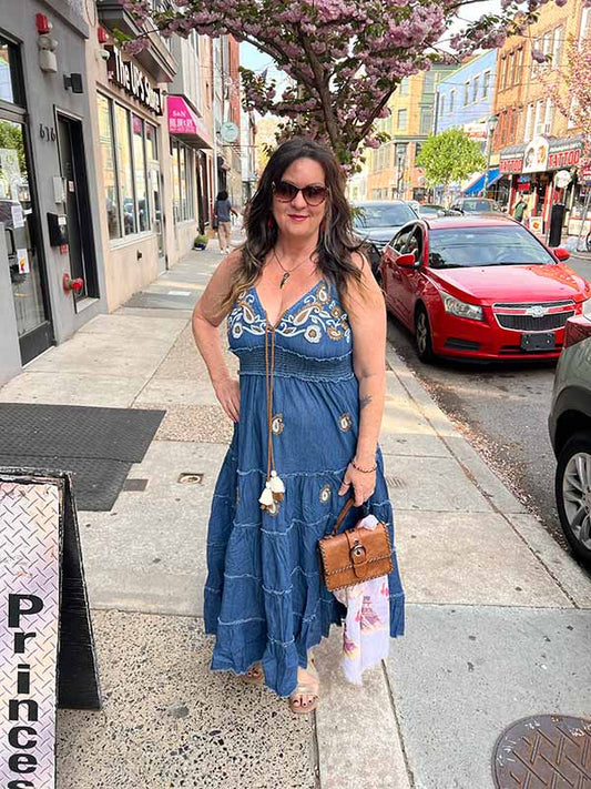 Owner Mary wearing a dark denim beaded maxi dress with adjustable straps on a city street with cherry blossoms