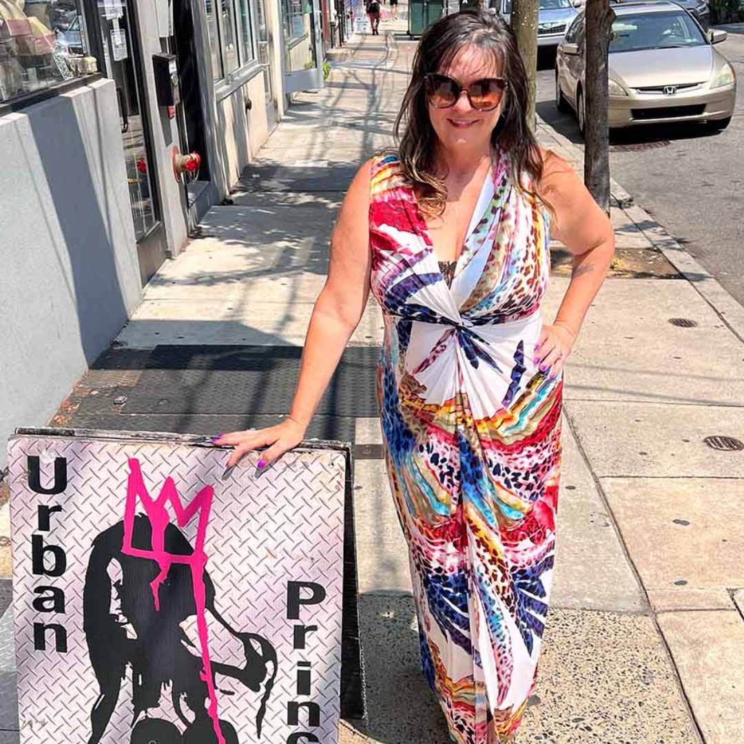 Model wearing white plunging v-dress with floral print, one hand on hip other on Urban Princess standing sign
