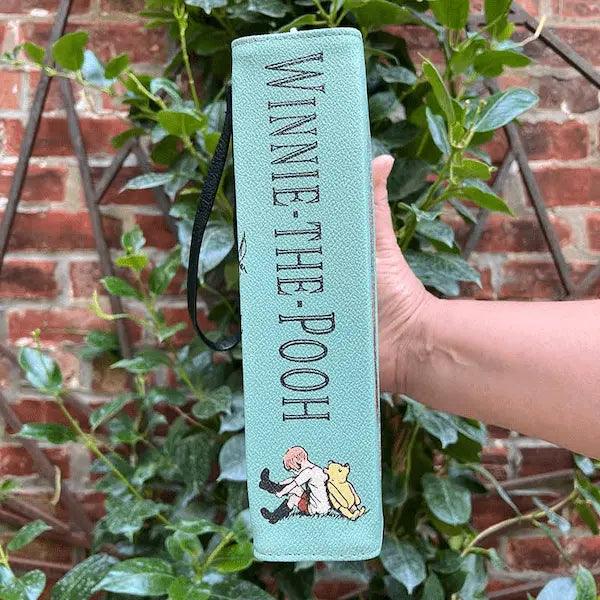 Light blue book with 'Winnie the Pooh' text held by a hand against a brick wall and greenery background. It is secretly a  storybook handbag.