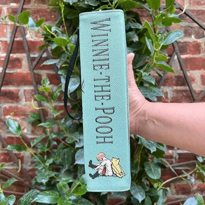 Light blue book with 'Winnie the Pooh' text held by a hand against a brick wall and greenery background. It is secretly a  storybook handbag.