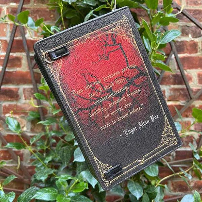 Decorative book with red cover and black text against a brick wall and green plants background. It is modeled after the book The Raven y Edgar Allen Poe and features and Edgar Allen Poe quote