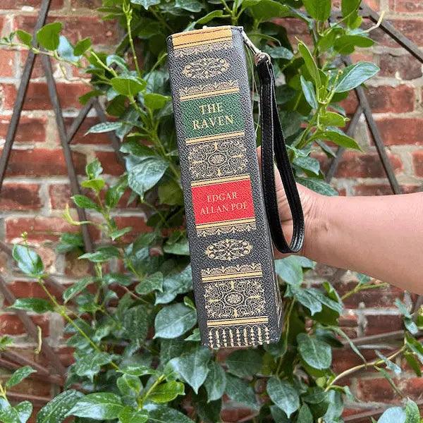 Another story book handbag - a book-shaped bag with 'The Raven' design held against a brick wall and green plants background. The Raven by Edgar Allen Poe