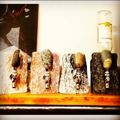4 Rock tap wine dispenser in different stone colors on a wooden shelf, far right dispenser has wine bottle displayed