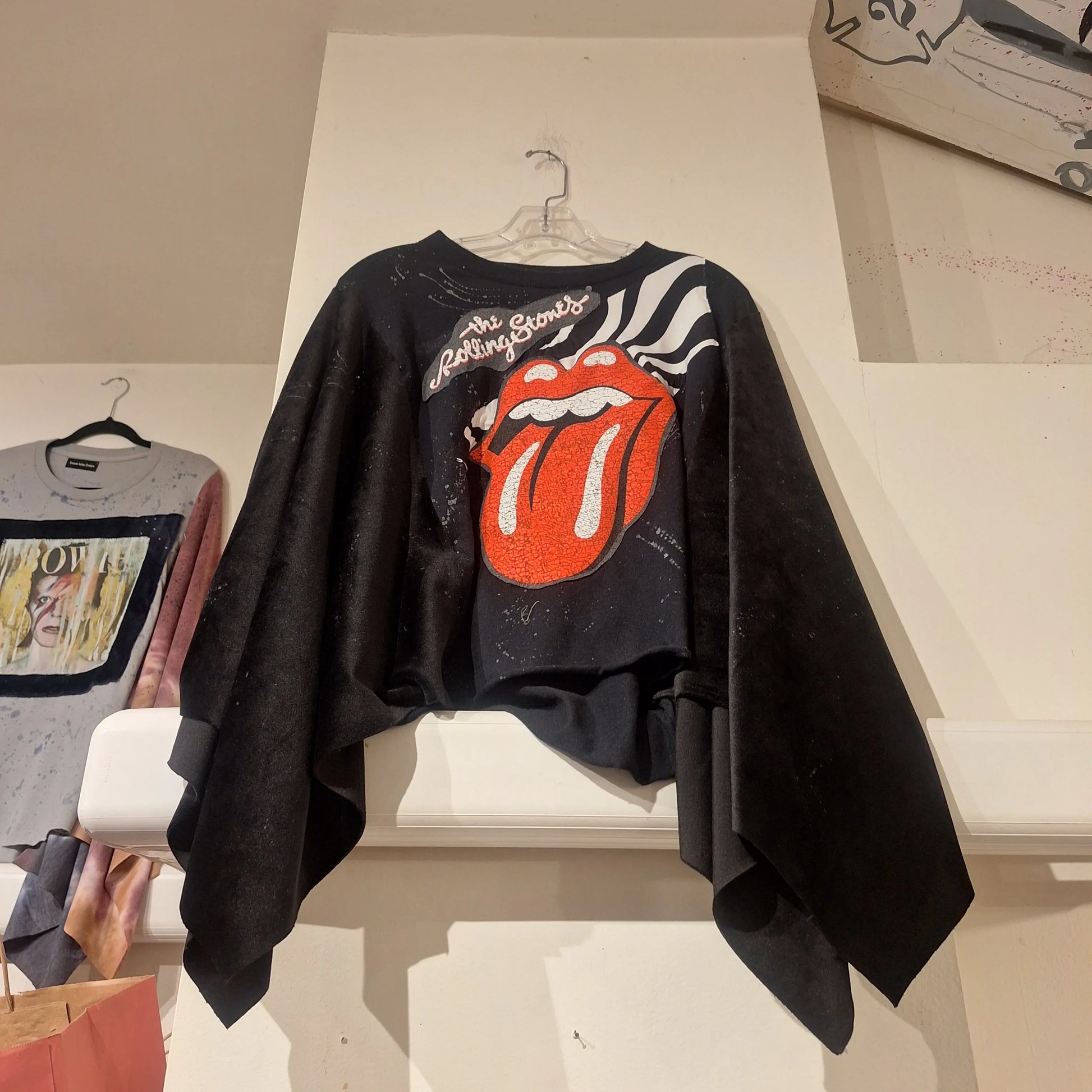 Custom Rolling Stones Poncho hanging on a wall. It is a black poncho with the Rolling Stones Tongue as an applique on the front.  In the Background there is a David Bowid Poncho hanging.