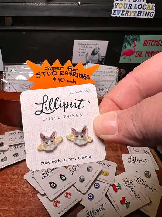 Hand holding a card with cat cosmo earrings labeled 'Lilliput Little Things' on a store shelf.