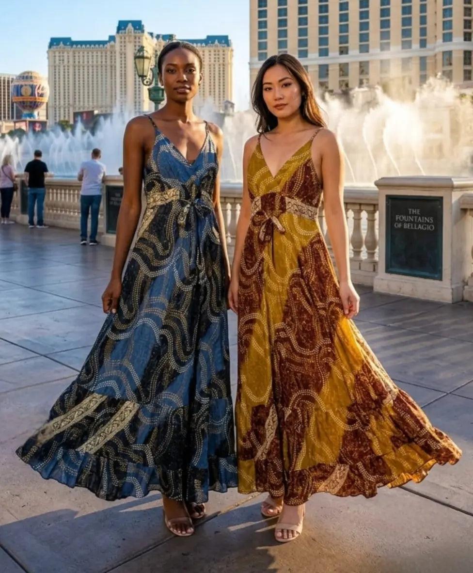 Two women wearing the Sari Maxi Dress in navy-gold swirl print and mustard-burgundy swirl print, posing at the Fountains of Bellagio in Las Vegas."