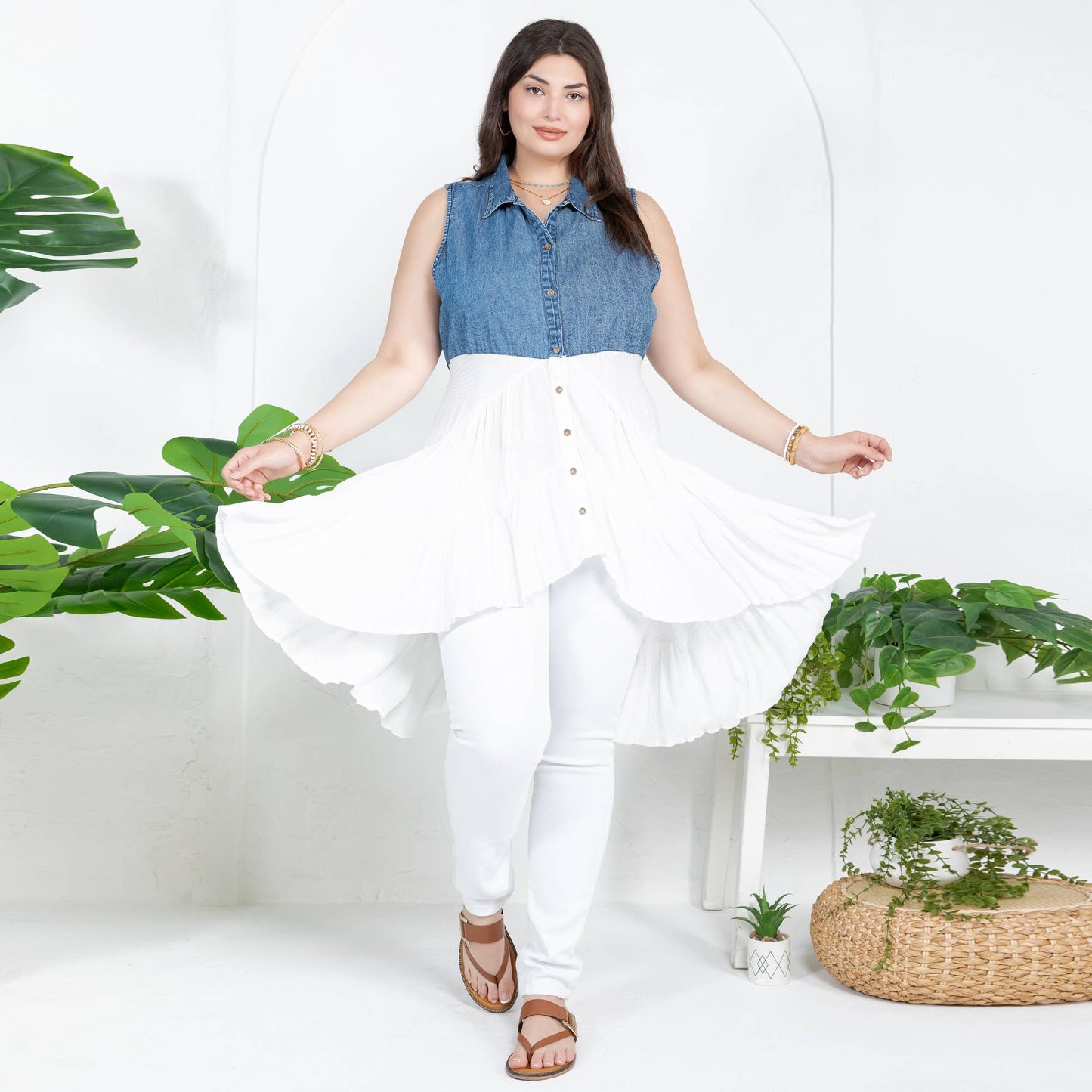 Woman wearing a  festival fit featuring  a tunic with a blue denim top and white skirt with plants in the background