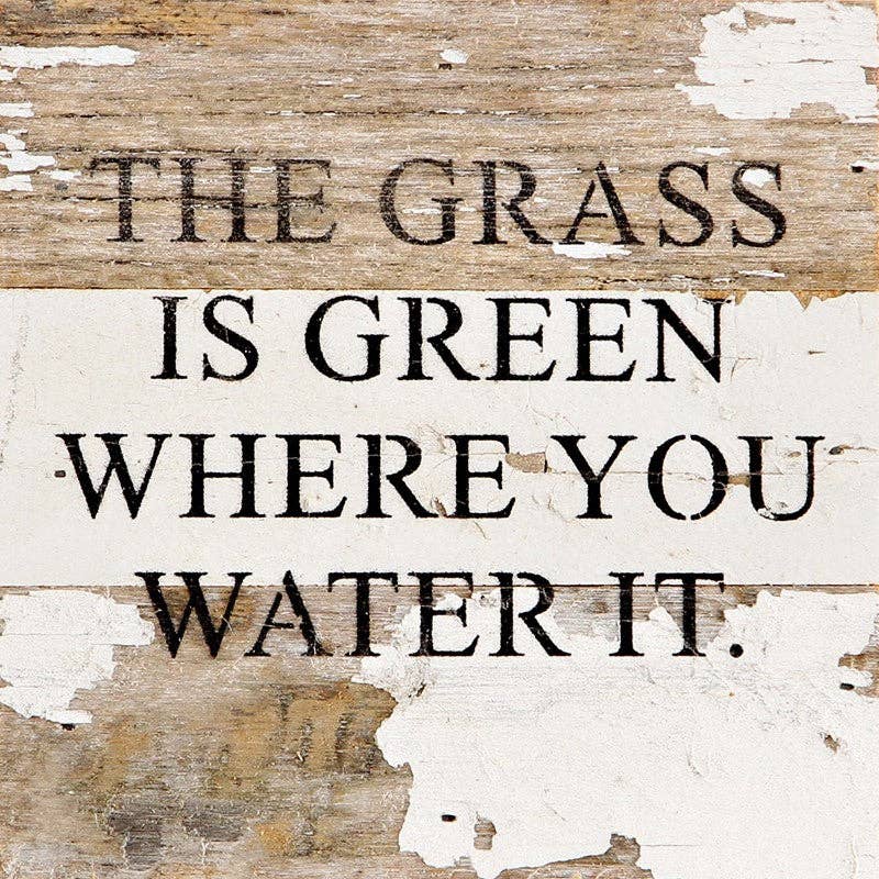 White distressed reclaimed wood sign with black text: “THE GRASS IS GREEN WHERE YOU WATER IT.”