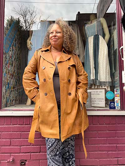 A woman with curly hair stands in front of a storefront window wearing a tan double-breasted leather coat over a dark top and patterned pants. The storefront has a pink brick wall and mannequins in the window display.