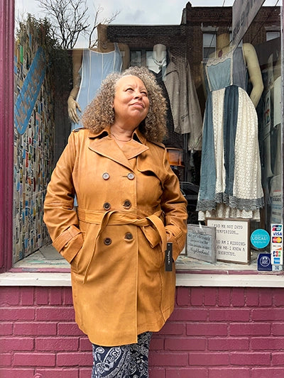 A woman with curly hair wearing a tan double-breasted leather coat stands in front of a shop window displaying dresses.