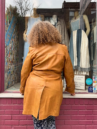 A person with curly light brown hair stands in front of a storefront window wearing a camel-brown leather trench coat. The window displays mannequins dressed in light blue and white dresses, set against a pink brick wall.