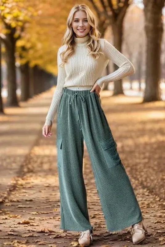Woman wearing a cream sweater and teal ribbed cargo pants standing on a leaf-covered path with trees in the background. Pants are soft enough to be pajamas