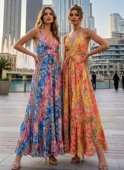 Two women wearing the Sari Maxi Dress in blue-pink tropical print and golden-orange floral print, standing in front of the Dubai Fountain with city skyline in the background.