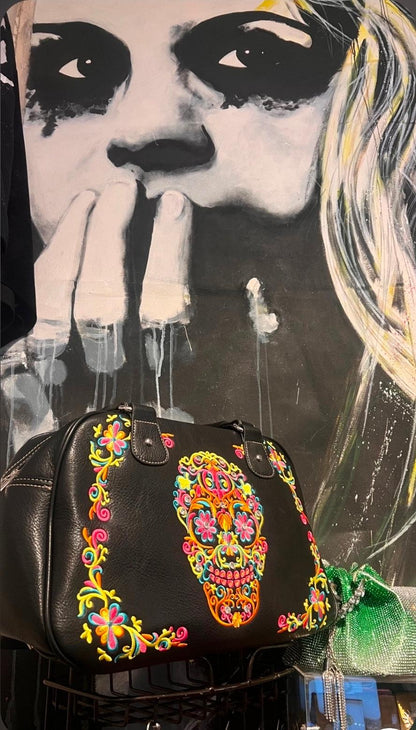 Black handbag with colorful skull design in front of a stylized portrait.
