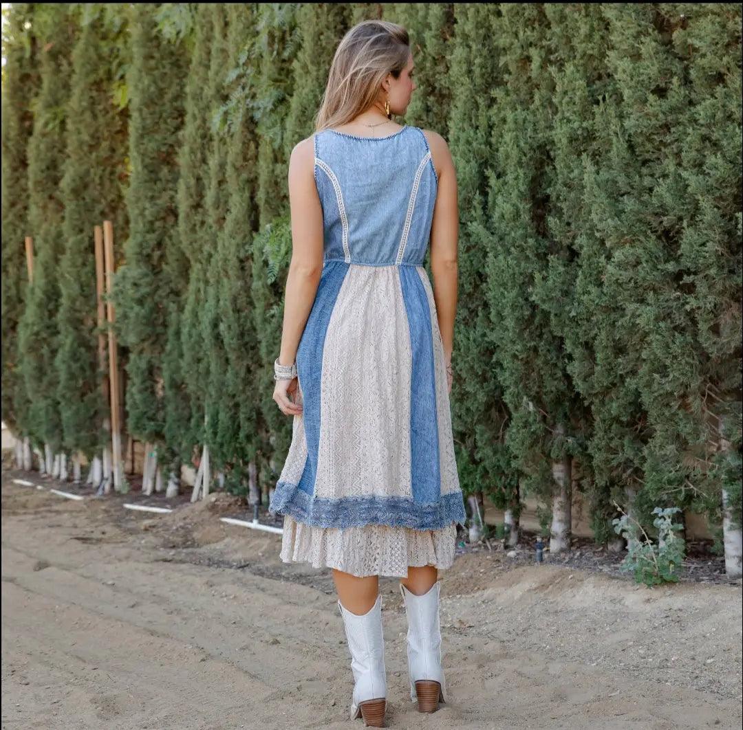 Woman wearing a blue and white denim and lace dress with boho vibes standing in front of a row of trees.