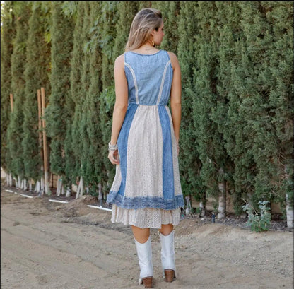 Woman wearing a blue and white denim and lace dress with boho vibes standing in front of a row of trees.
